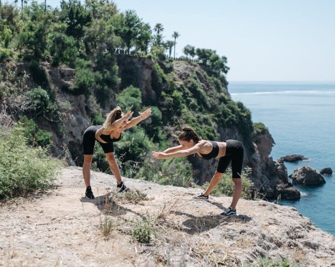 Two women in activewear stretching on a cliffside with an ocean view, embracing a healthy outdoor lifestyle.